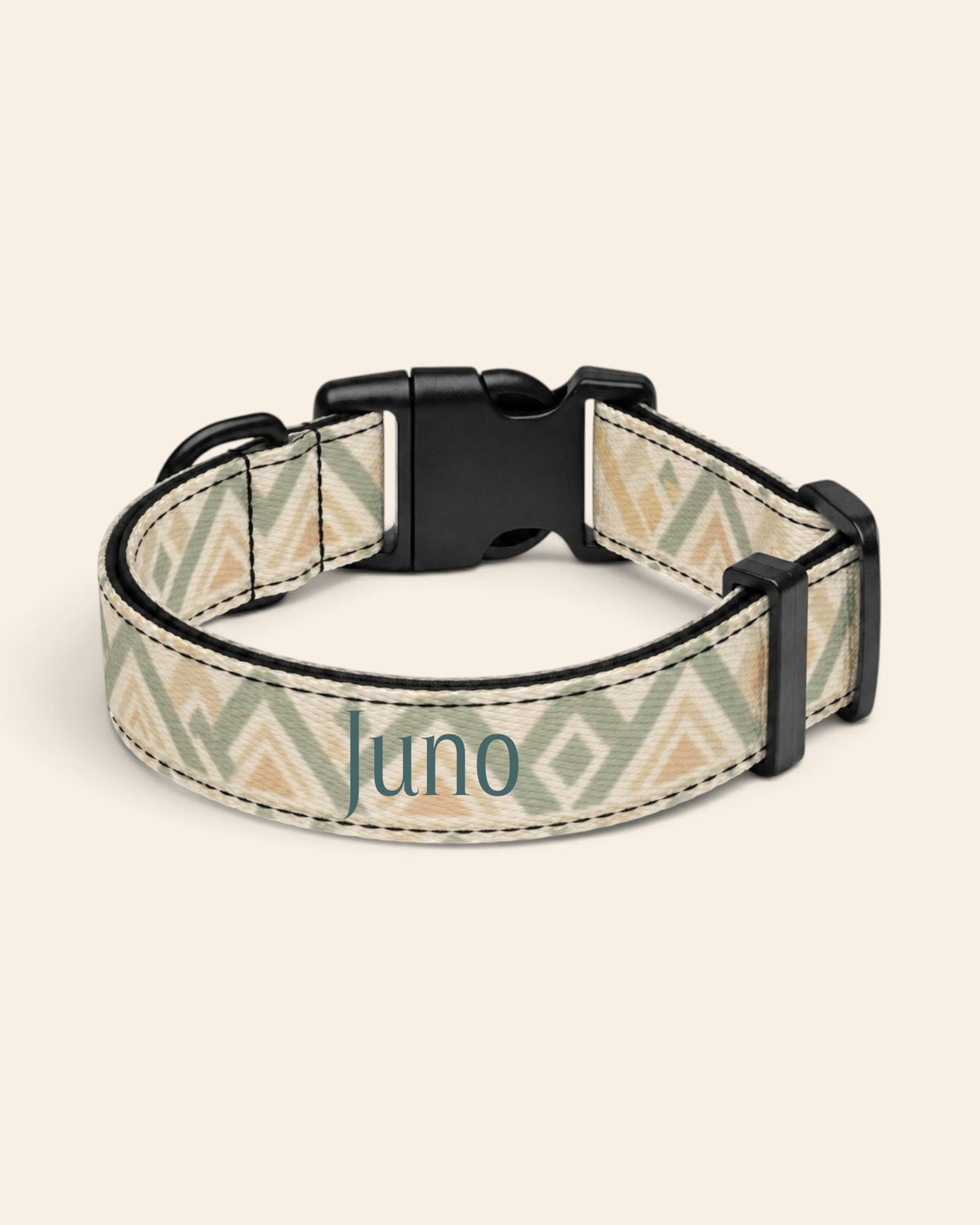 The collar alone on a plain cream background, lying in a loose loop that shows the printed webbing, a black side-release buckle and a black slider. The strap is cream, banded with sage and apricot chevrons and diamond geometric motifs, with the sample name Juno printed along it.