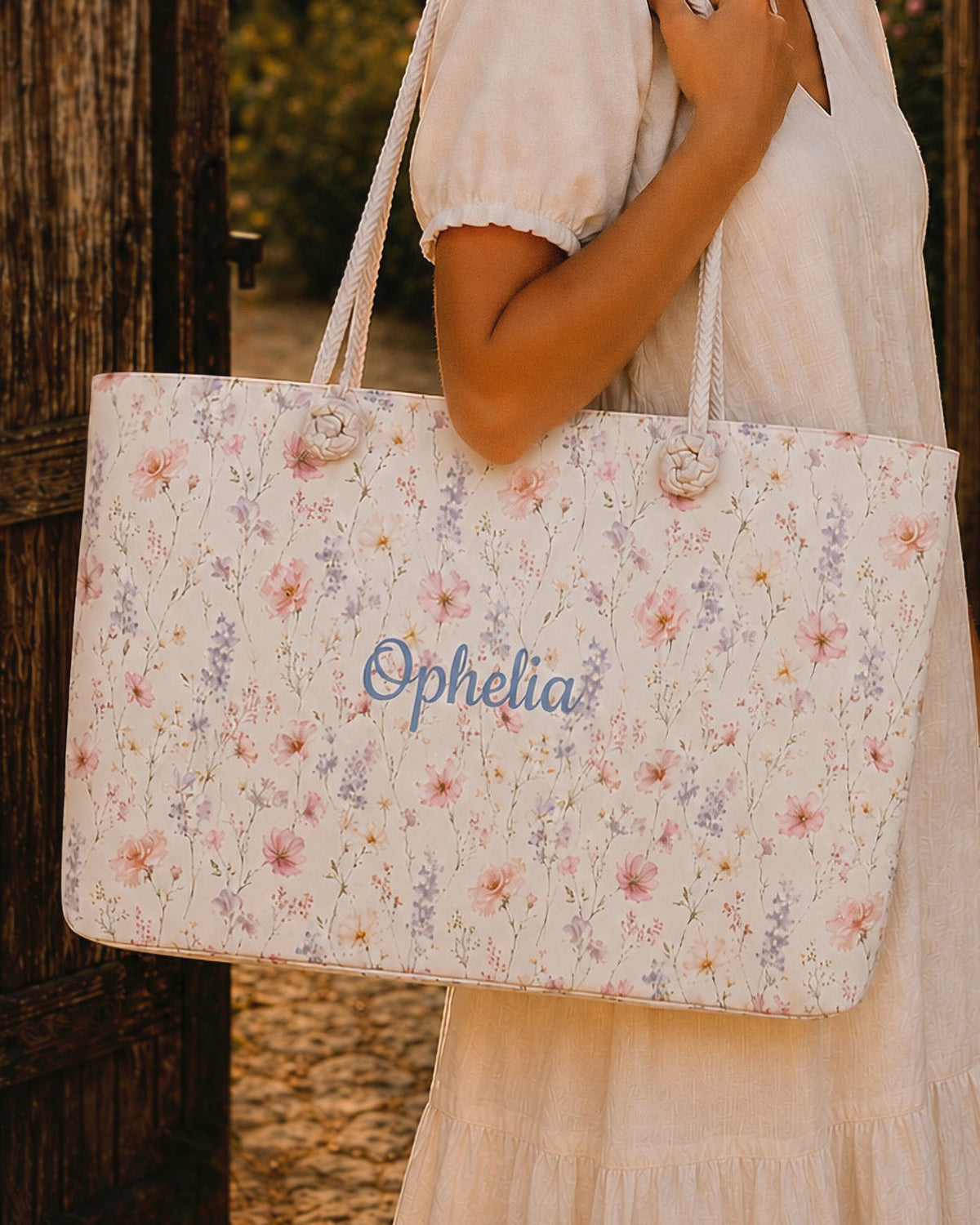 A woman carries the weekender tote on her shoulder, wearing a cream linen dress, against a weathered wood plank wall. The bag is white, densely printed with pink, lilac and apricot blooms, with the sample name Ophelia in a flowing script across the side panel and thick cream rope handles knotted through the top corners.
