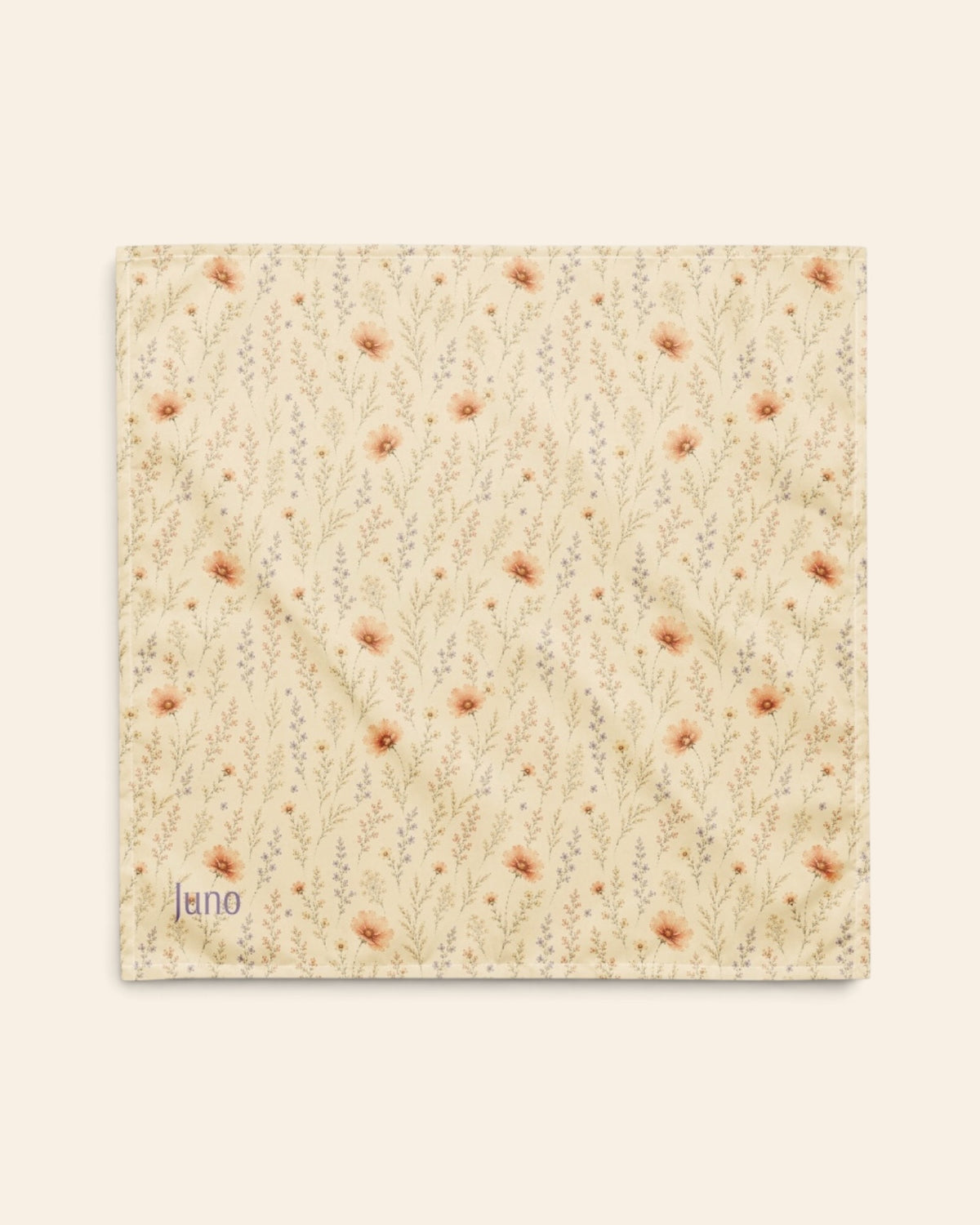 The bandana laid flat as a full square on a cream background, the print running on the diagonal. It is soft butter-cream, printed with small apricot and coral blooms among fine sage stems, with the sample name Juno printed in the lower left corner.