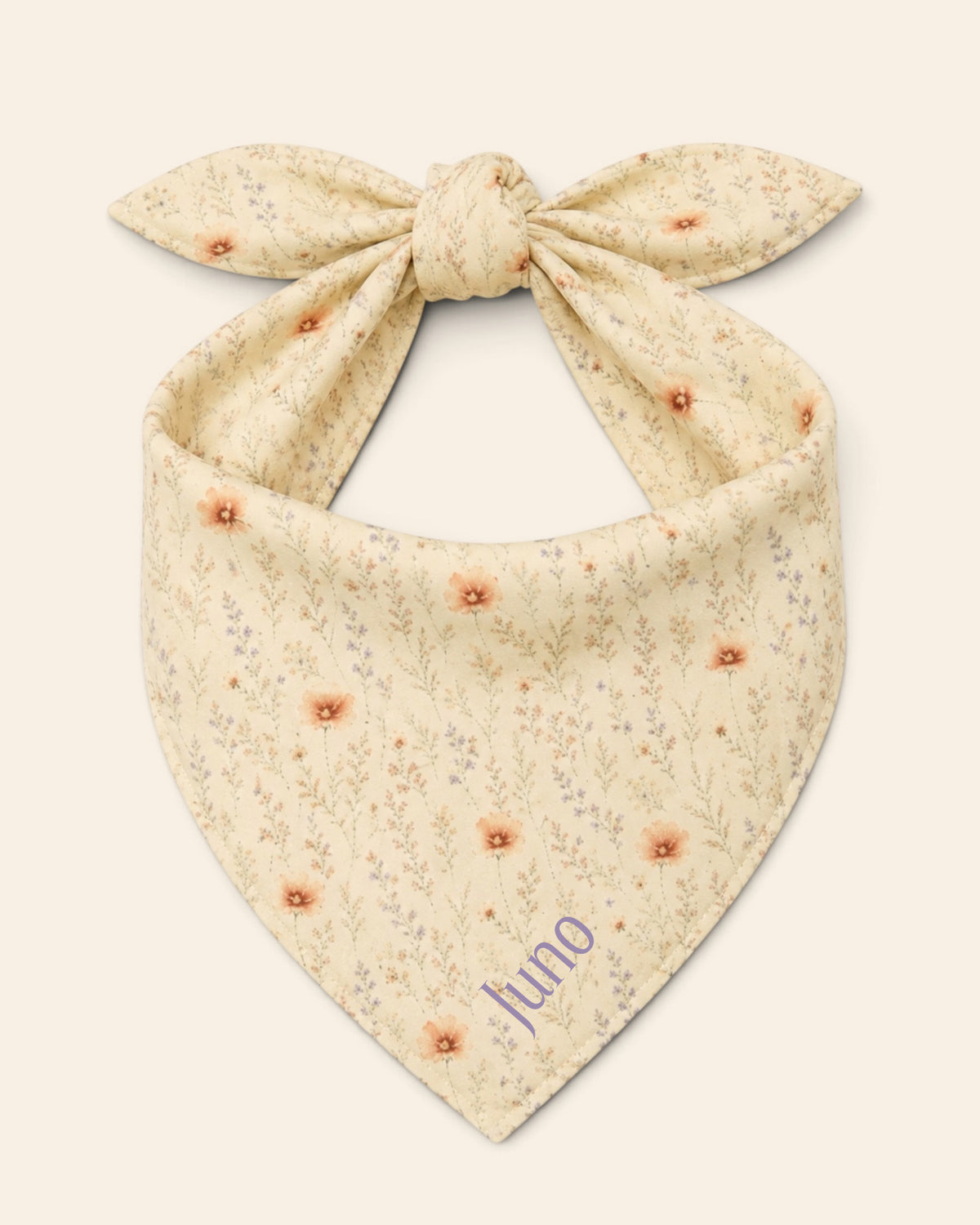 The bandana alone on a plain cream background, knotted so the two tail ends stand up and the triangle hangs below. It is soft butter-cream, printed with small apricot and coral blooms among fine sage stems, with the sample name Juno printed on the lower right panel.
