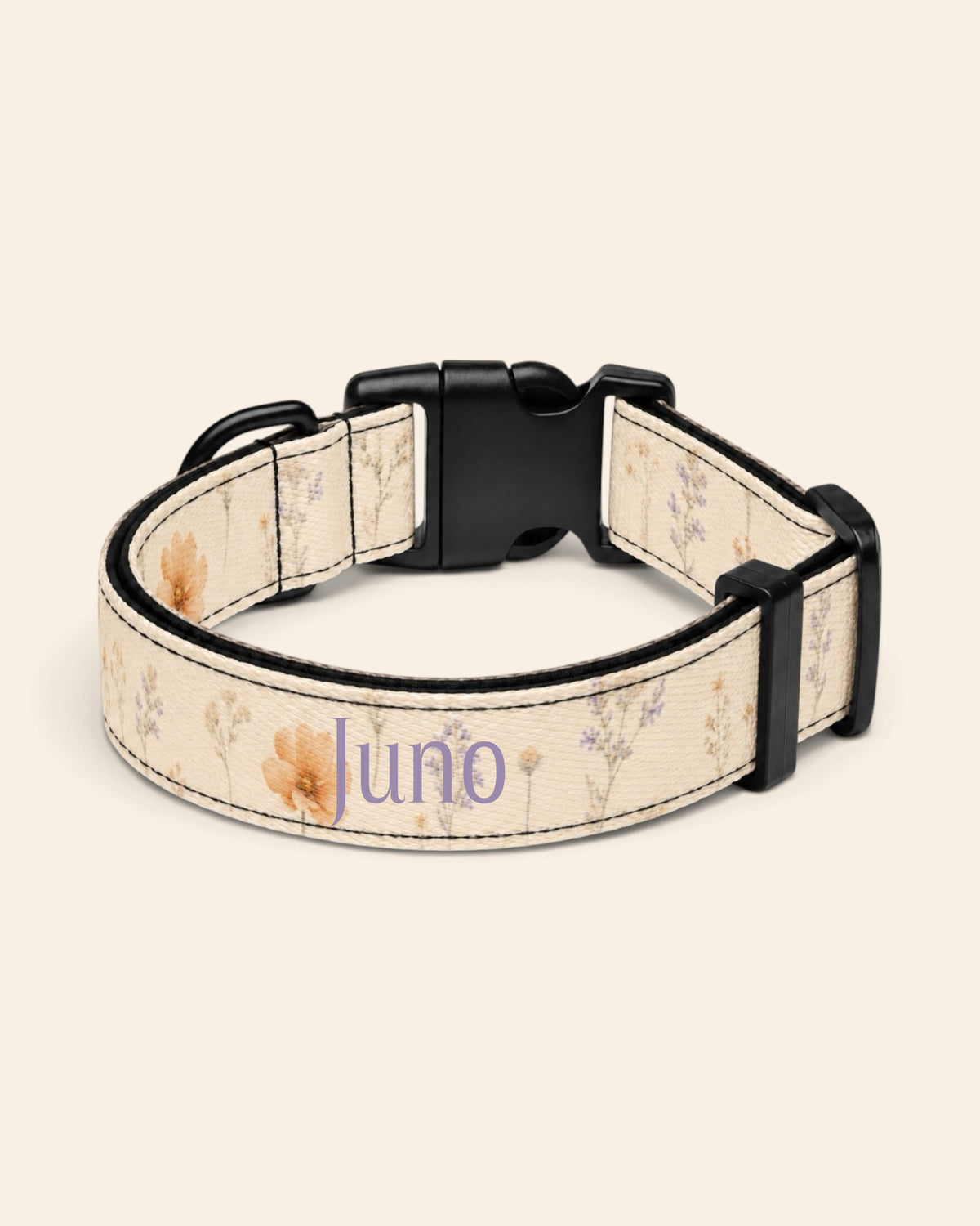 The collar alone on a plain cream background, lying in a loose loop that shows the printed webbing, a black side-release buckle and a black slider. The strap is soft butter-cream, printed with small apricot and coral blooms among fine sage stems, with the sample name Juno printed along it.