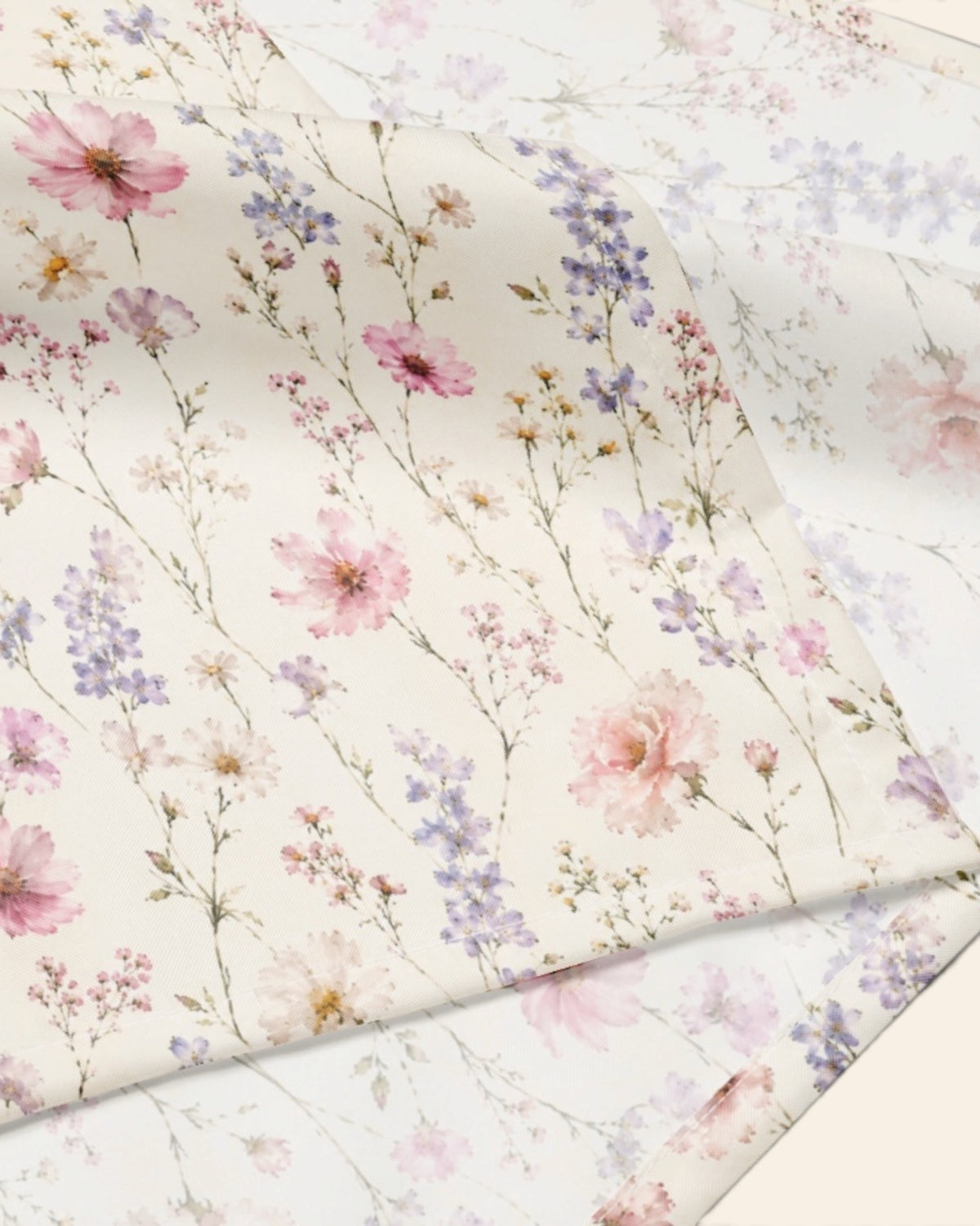 A close crop of the bandana fabric with one corner folded back over a soft cream ground, showing the print at full scale - white, densely printed with pink, lilac and periwinkle wildflowers.