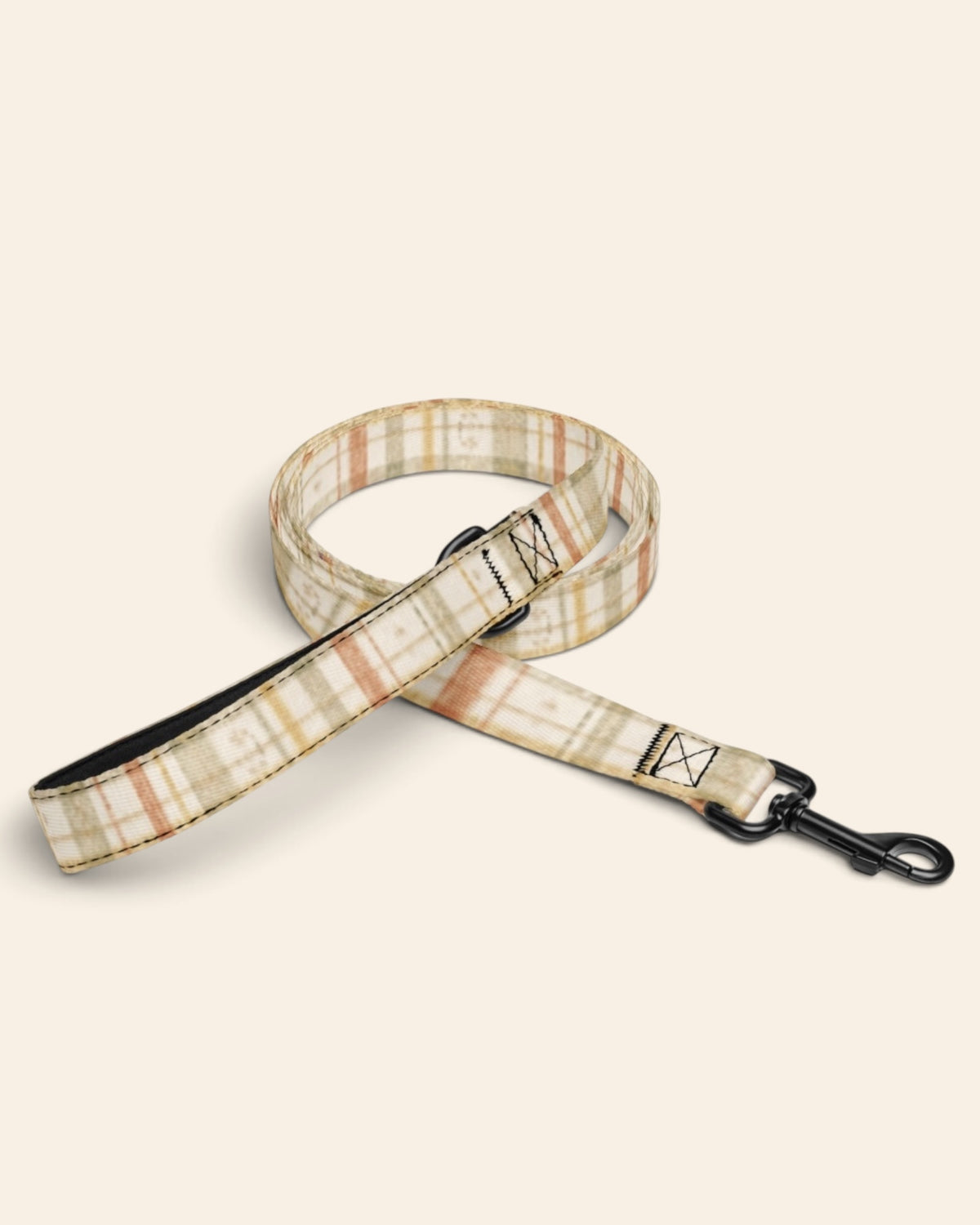 The full length of the leash hangs from a tree branch outdoors, the padded handle loop at the top and the black swivel clasp at the bottom, conifers and a mountain slope beyond in golden light. The webbing is a sage, gold and apricot plaid on cream, banded with rows of small motifs, and the sample name Juno reads down the handle.