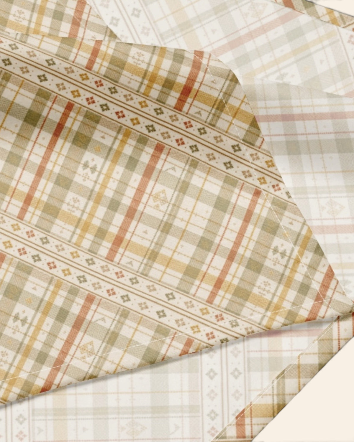 A close crop of the bandana fabric with one corner folded back over a soft cream ground, showing the print at full scale - a sage, gold and apricot plaid on cream, banded with rows of small motifs.
