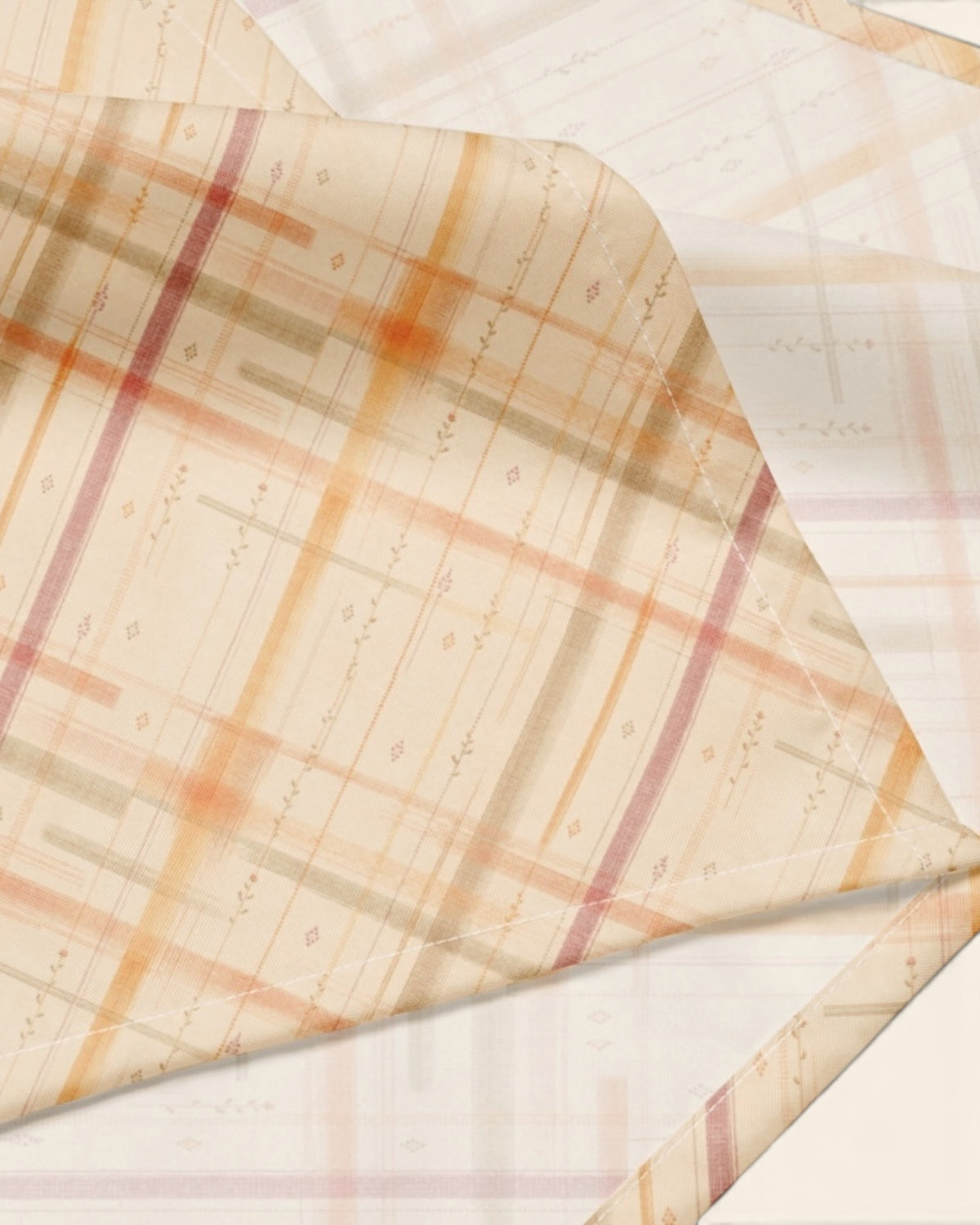 A close crop of the bandana fabric with one corner folded back over a soft cream ground, showing the print at full scale - a cream plaid crossed by apricot, rose and sage lines.