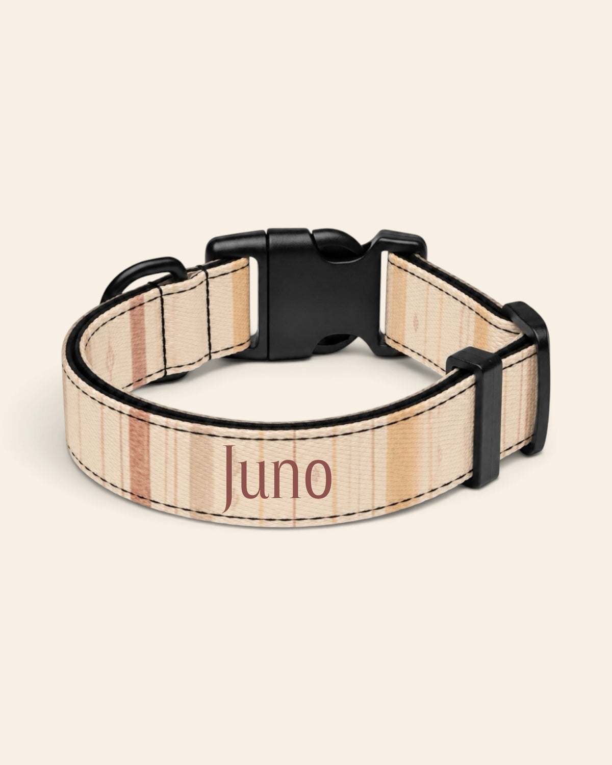 The collar alone on a plain cream background, lying in a loose loop that shows the printed webbing, a black side-release buckle and a black slider. The strap is a cream plaid crossed by apricot, rose and sage lines, with the sample name Juno printed along it.