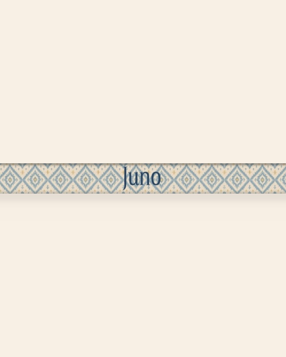 A close, straight-on crop of the leash strap laid flat on a cream background, showing the print and the sample name Juno running along the webbing. The strap is cream, printed with dusty blue ikat diamonds picked out with gold and small cross motifs.