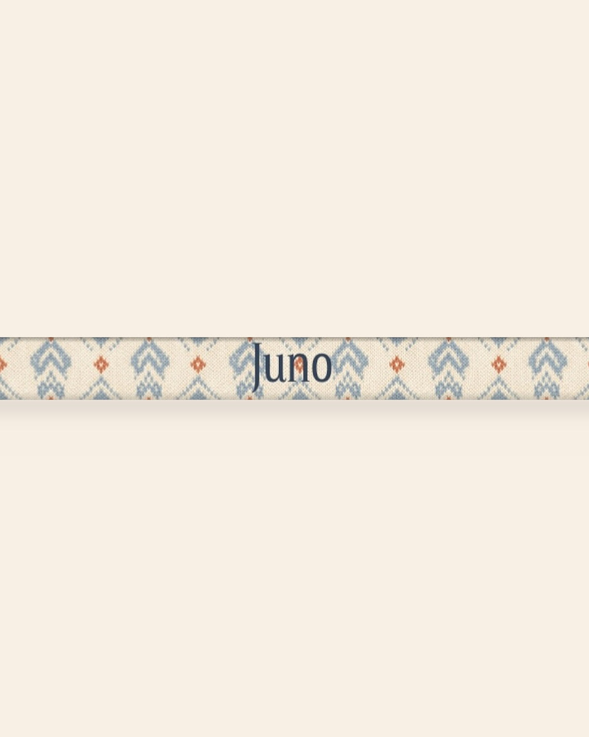 A close, straight-on crop of the leash strap laid flat on a cream background, showing the print and the sample name Juno running along the webbing. The strap is cream, printed with dusty blue zigzag chevrons and stepped diamond motifs with small rust accents.